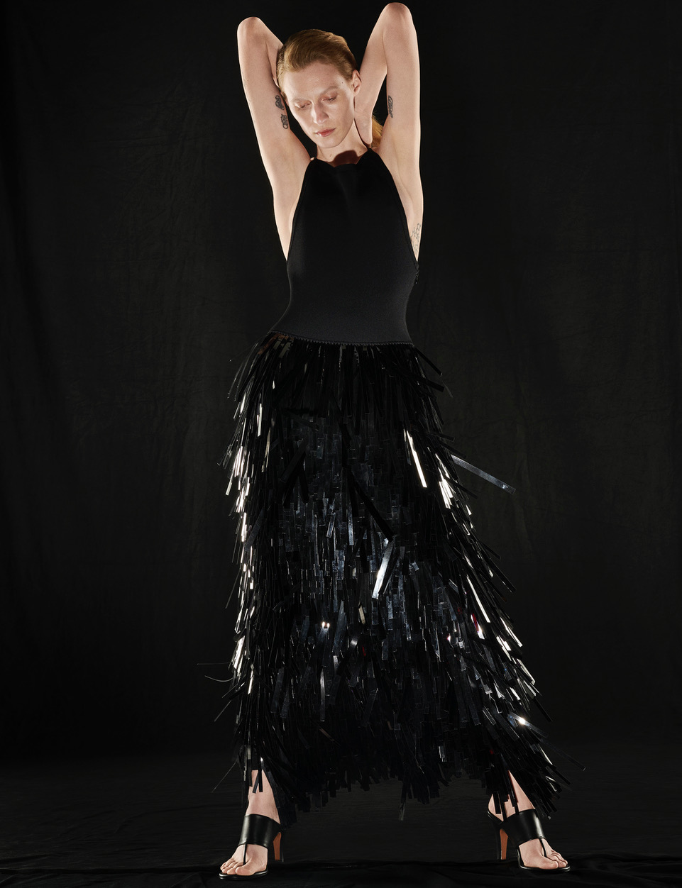 Elegant woman poses in a black halter dress with fringed skirt against a dark backdrop in a fashion photo showcasing sleek style.