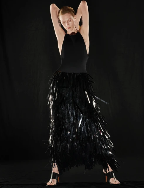 Elegant woman poses in a black halter dress with fringed skirt against a dark backdrop in a fashion photo showcasing sleek style.