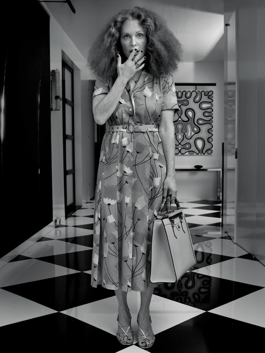 A black and white shot of an adult female in a floral dress standing on a checkered floor smoking a cigarette while holding a purse.