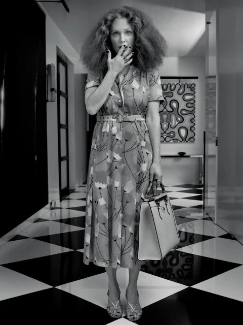 A black and white shot of an adult female in a floral dress standing on a checkered floor smoking a cigarette while holding a purse.