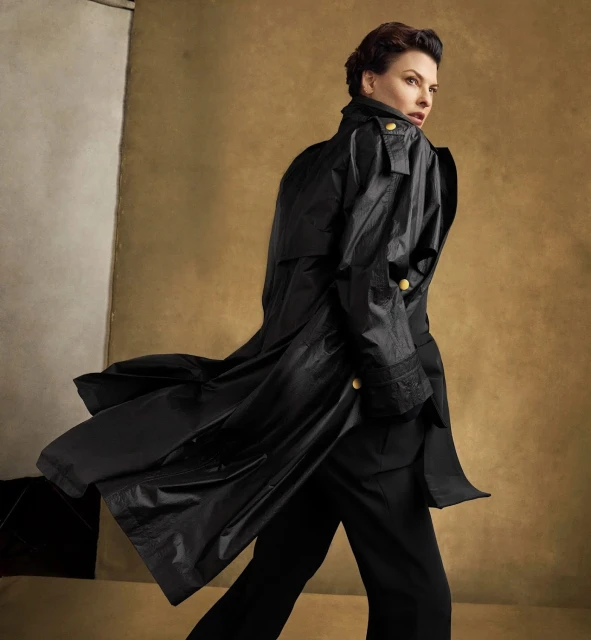 Brunette adult woman in a long black coat turns over her shoulder against a tan and white mottled studio backdrop with soft lighting.