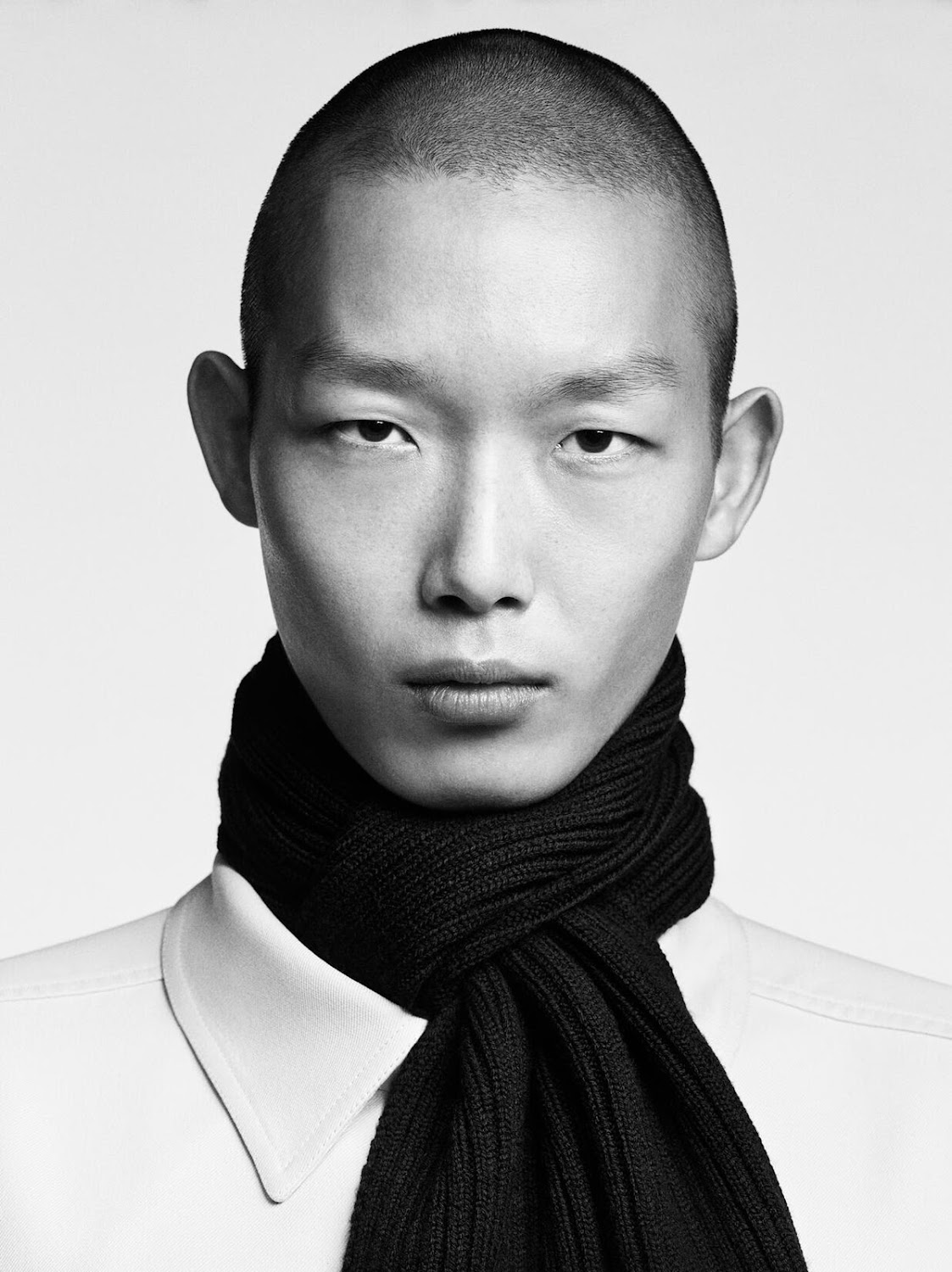 Close-up portrait of a male model with a shaved head wearing a white shirt and a black scarf against a seamless white background.