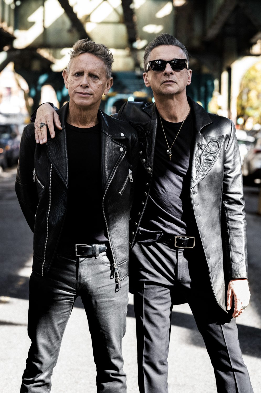 Two adult males stand on a city street under an overpass, dressed in leather jackets and dark clothing. They wear sunglasses and belts.