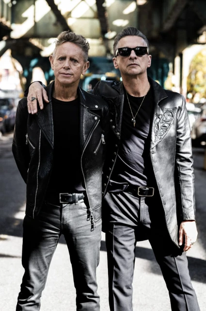 Two adult males stand on a city street under an overpass, dressed in leather jackets and dark clothing. They wear sunglasses and belts.
