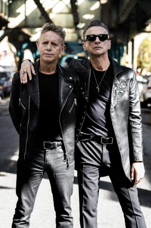 Two adult males stand on a city street under an overpass, dressed in leather jackets and dark clothing. They wear sunglasses and belts.