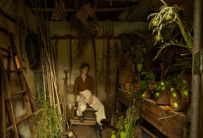 Person in brown shirt and light trousers sitting inside a rustic shed filled with garden tools and plants