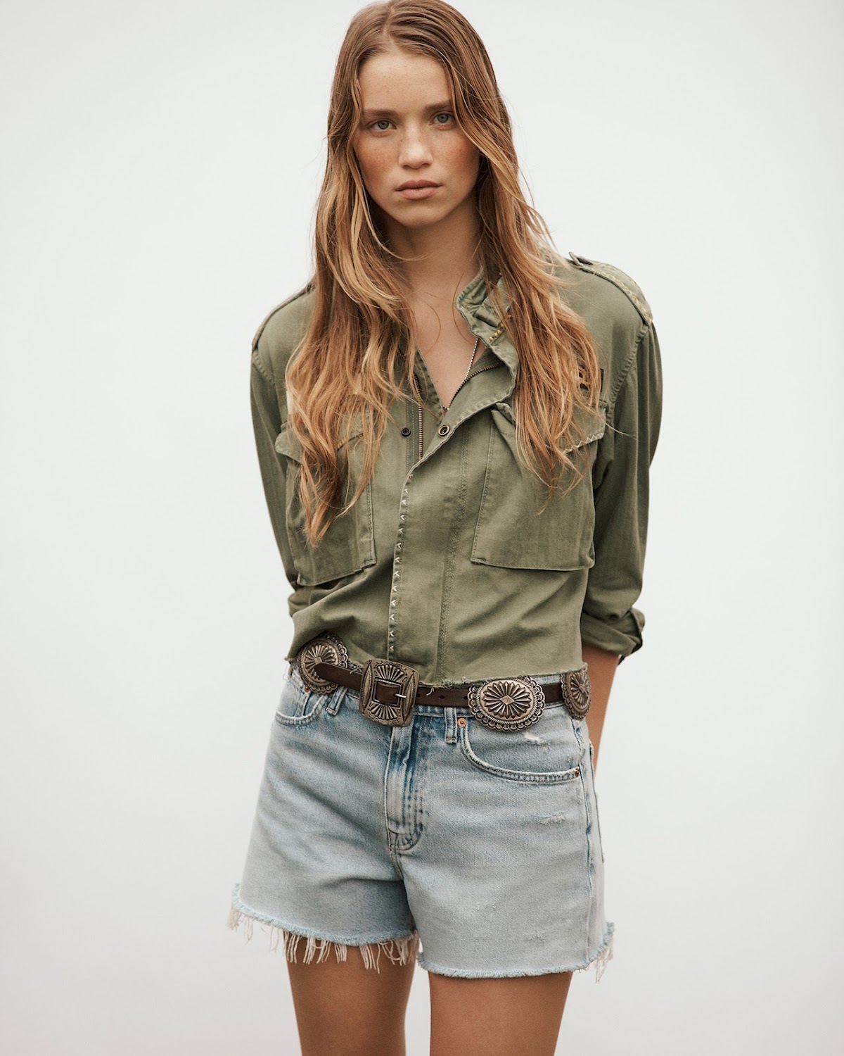 A woman with long brown hair poses in an olive button-down jacket and denim shorts with a concho belt against a light background.