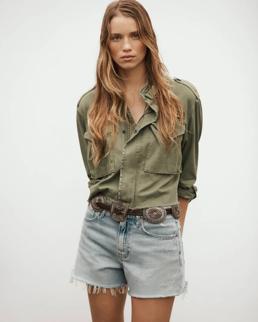 A woman with long brown hair poses in an olive button-down jacket and denim shorts with a concho belt against a light background.