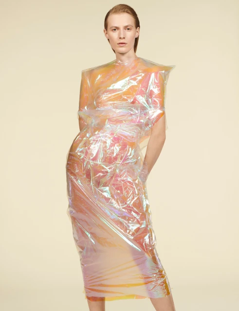 Female fashion model poses in a futuristic, semi-transparent plastic dress against a neutral backdrop in a studio setting.