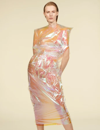Female fashion model poses in a futuristic, semi-transparent plastic dress against a neutral backdrop in a studio setting.