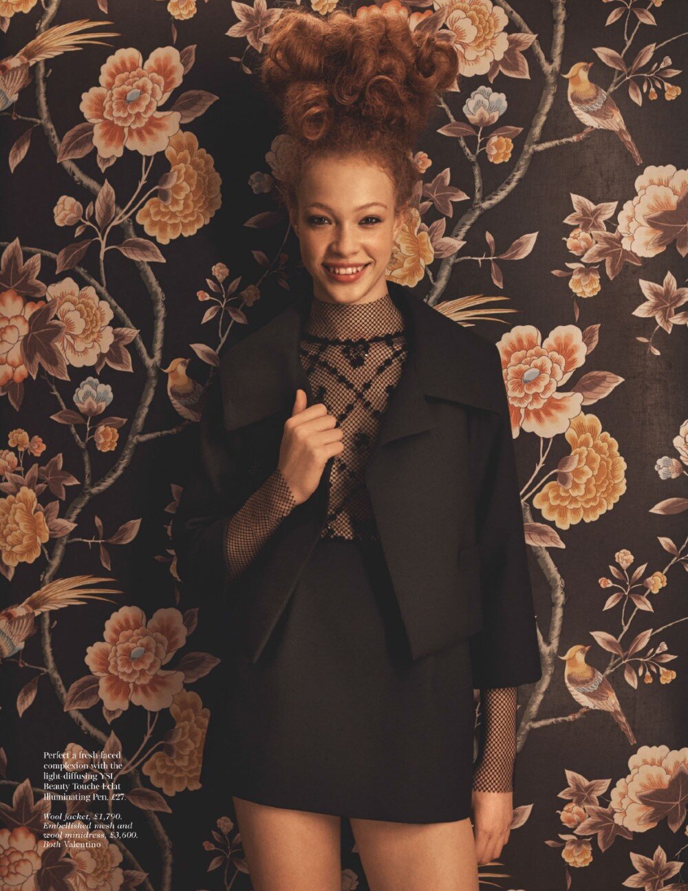 Portrait of a smiling woman with voluminous red hair wearing a black jacket against a dark floral wallpaper background.