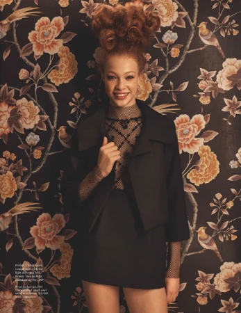 Portrait of a smiling woman with voluminous red hair wearing a black jacket against a dark floral wallpaper background.
