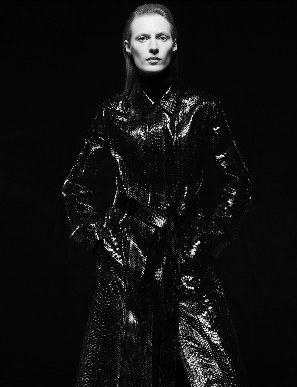 Model in glossy black textured coat with belt against a dark background featuring make-up by Francelle Daly