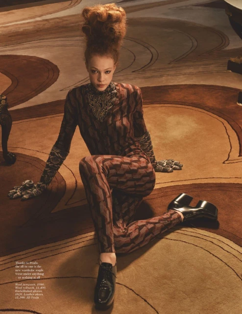 A female fashion model with a high auburn beehive hairstyle poses in a patterned jumpsuit and platform shoes on a patterned carpet.