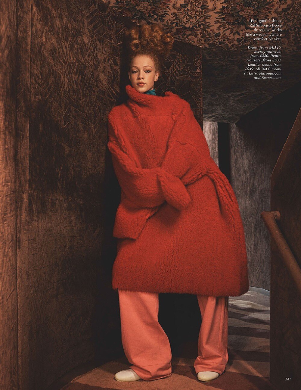 A red-haired woman in a bold red coat and loose pink pants poses in a dimly lit, patterned hallway, creating a striking fashion statement.