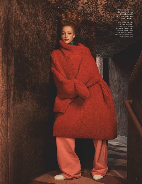 A red-haired woman in a bold red coat and loose pink pants poses in a dimly lit, patterned hallway, creating a striking fashion statement.