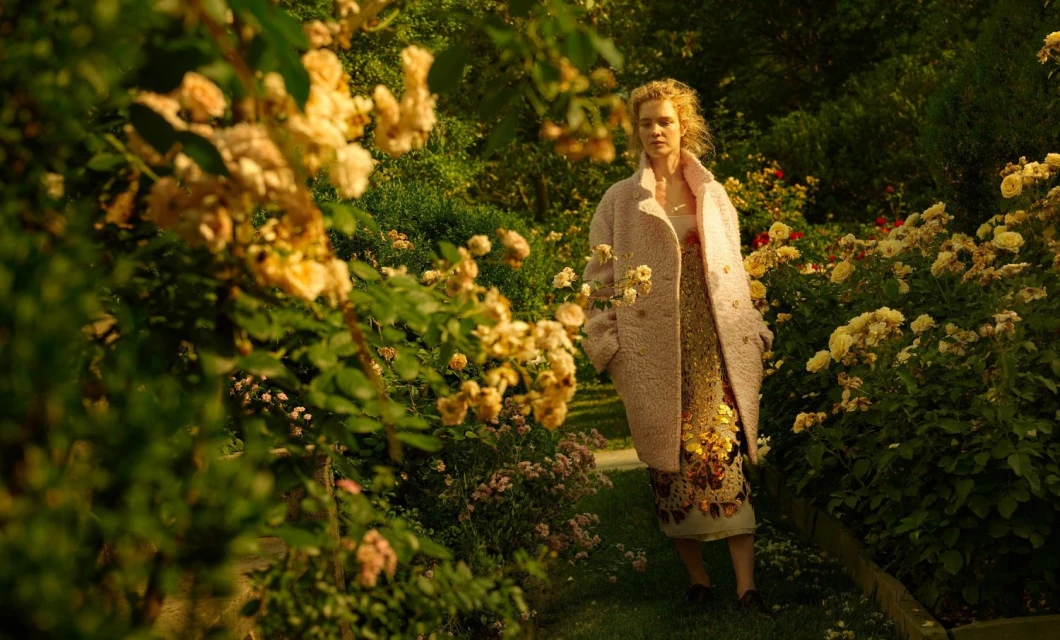 A woman in a pink coat stands in a rose garden, surrounded by yellow roses and greenery. She wears a gold patterned dress in natural light.