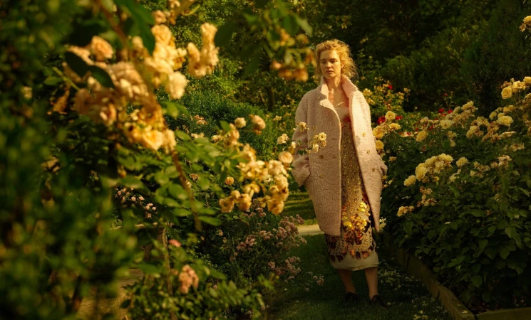 A woman in a pink coat stands in a rose garden, surrounded by yellow roses and greenery. She wears a gold patterned dress in natural light.