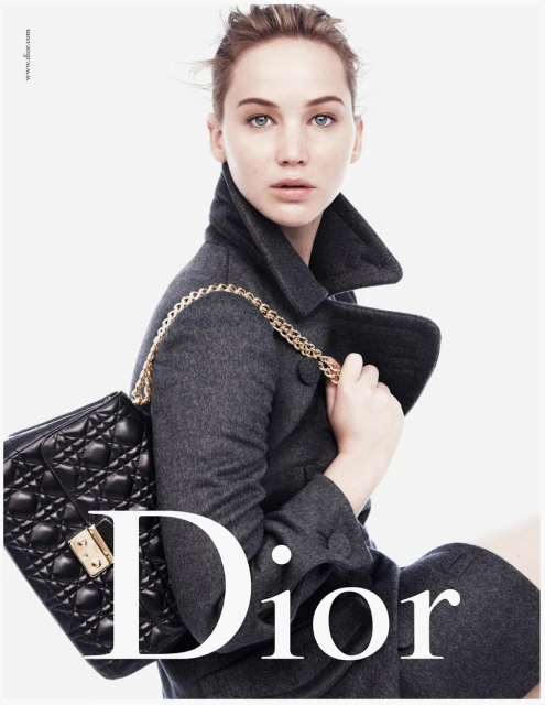 Jennifer Lawrence poses with a black quilted Dior bag and grey wool coat against a white background in a fashion editorial shot.