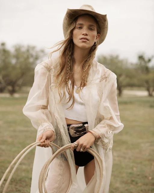 A woman in a cowboy hat and sheer robe holds a lasso in an outdoor field, styled with a Western belt and braided hair accents.