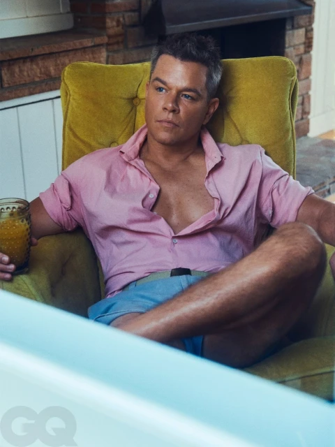 Matt Damon sits in a yellow armchair wearing a pink shirt and blue shorts, holding a beverage. GQ logo visible in the corner.