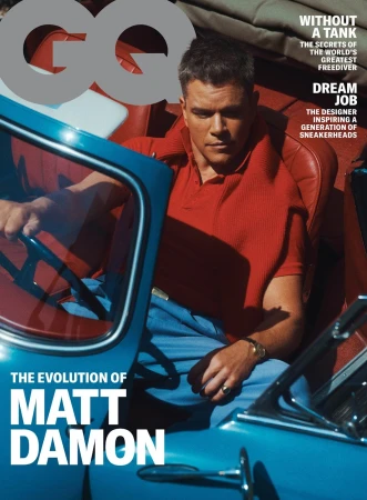 Matt Damon poses in a blue vintage car for the cover of GQ magazine. He wears a red collared shirt and light blue pants.