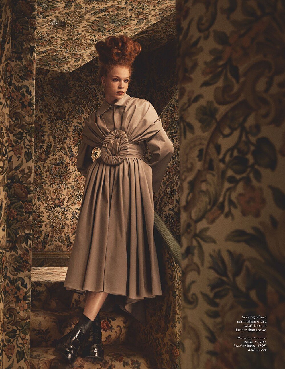 Fashion editorial photo of a woman in a draped coat dress and black boots posing in a wallpapered interior with a unique hairstyle.