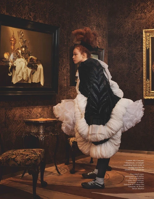 Fashion shot of a young woman in an elaborate black and white dress posing indoors in a room with paintings on patterned wallpaper.