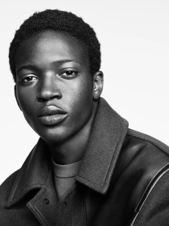 Close-up portrait of a young Black man wearing a dark collared jacket against a plain white background.