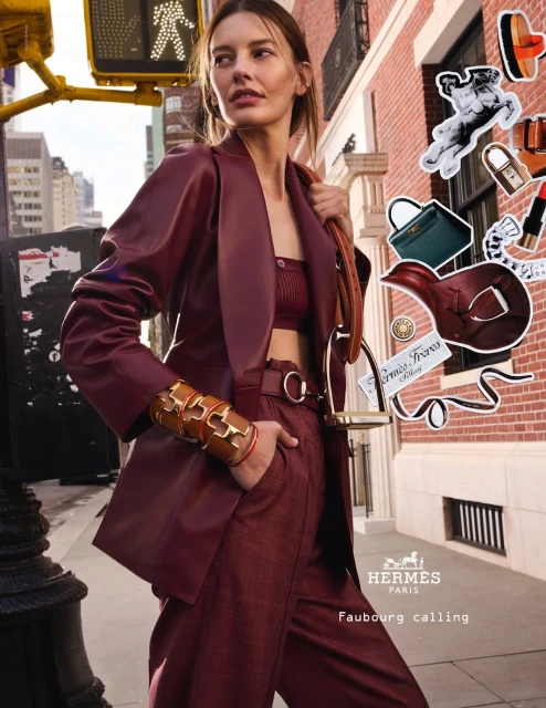 Fashionable woman in maroon pantsuit and crop top poses in an urban setting with a crosswalk and architectural details, wearing gold bracelets.