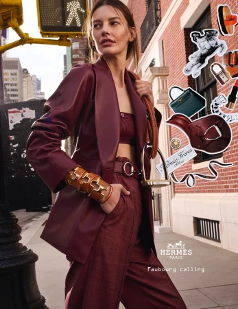 Fashionable woman in maroon pantsuit and crop top poses in an urban setting with a crosswalk and architectural details, wearing gold bracelets.