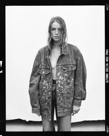 Black and white portrait of a young adult woman wearing a denim jacket with paint splatters, standing in front of a white backdrop.