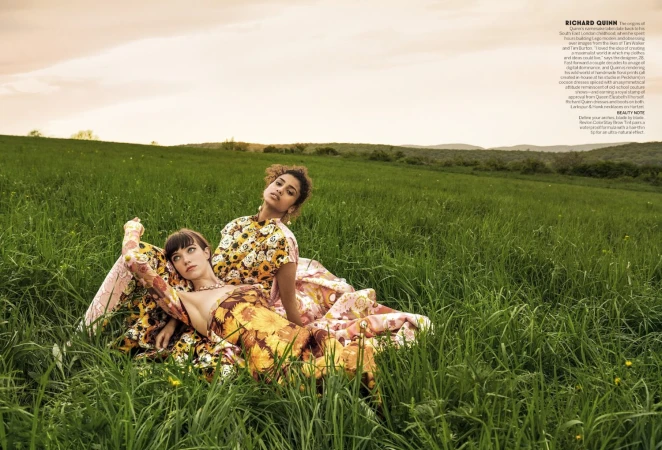 Two female models in patterned floral dresses reclining in a lush green meadow under an overcast sky.