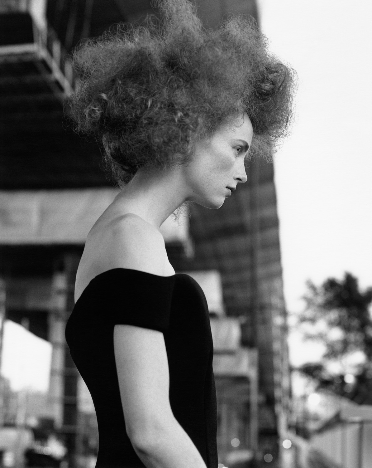 Black and white portrait of a woman with afro-textured hair wearing an off-the-shoulder dress against a blurred background of urban structures.