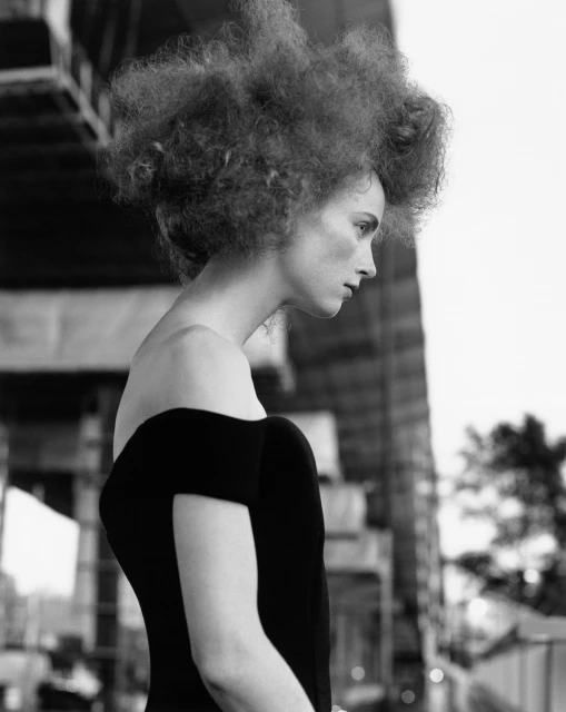 Black and white portrait of a woman with afro-textured hair wearing an off-the-shoulder dress against a blurred background of urban structures.
