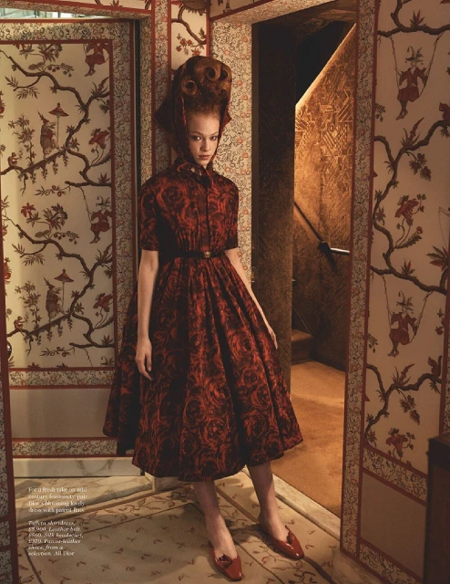 A redhead model in a red floral dress stands in a wallpapered corridor with red shoes, a red belt, and a high coiffure hairstyle.