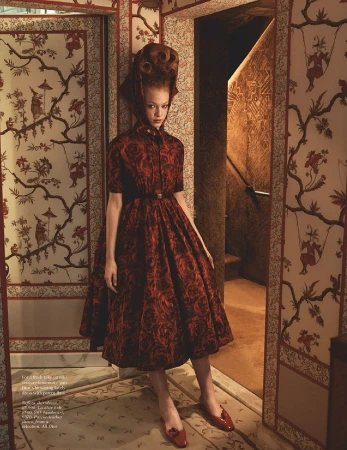 A redhead model in a red floral dress stands in a wallpapered corridor with red shoes, a red belt, and a high coiffure hairstyle.