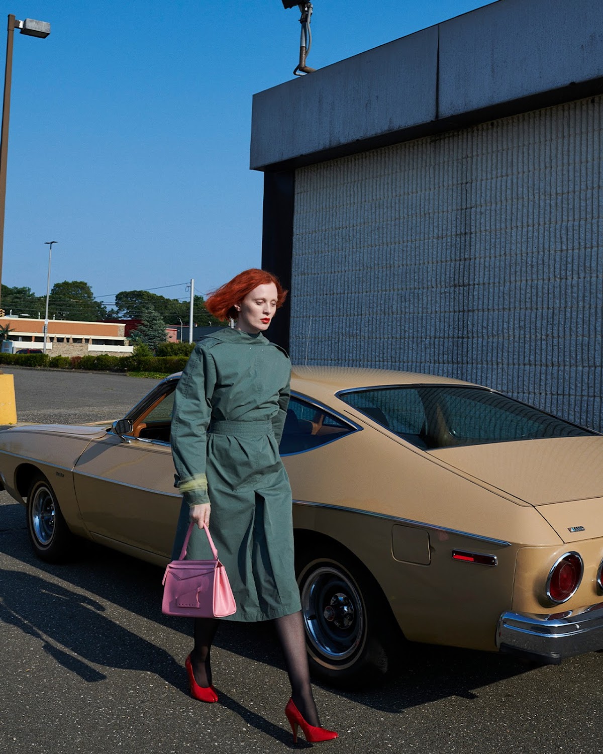 Elegant red-haired woman in green dress, red heels, and pink purse walking by a tan classic car near a concrete building.