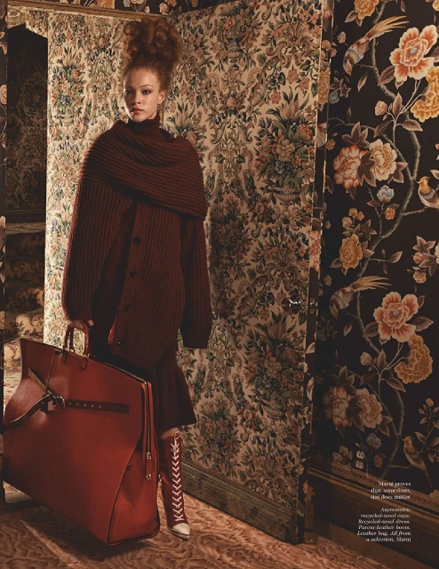 Fashionable woman with a red suitcase in a room with floral wallpaper, wearing a red knitted cardigan, red skirt, and lace-up boots.