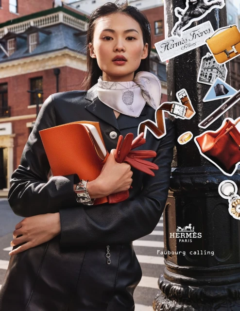 Elegant Asian woman in a black leather suit holds an orange document case and gloves, leaning against a lamp post in an urban setting.