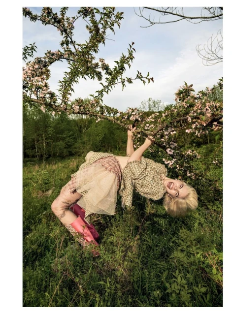 Woman in a patterned jacket and pink boots hangs upside down from a blossoming tree in a grassy field on a sunny day.