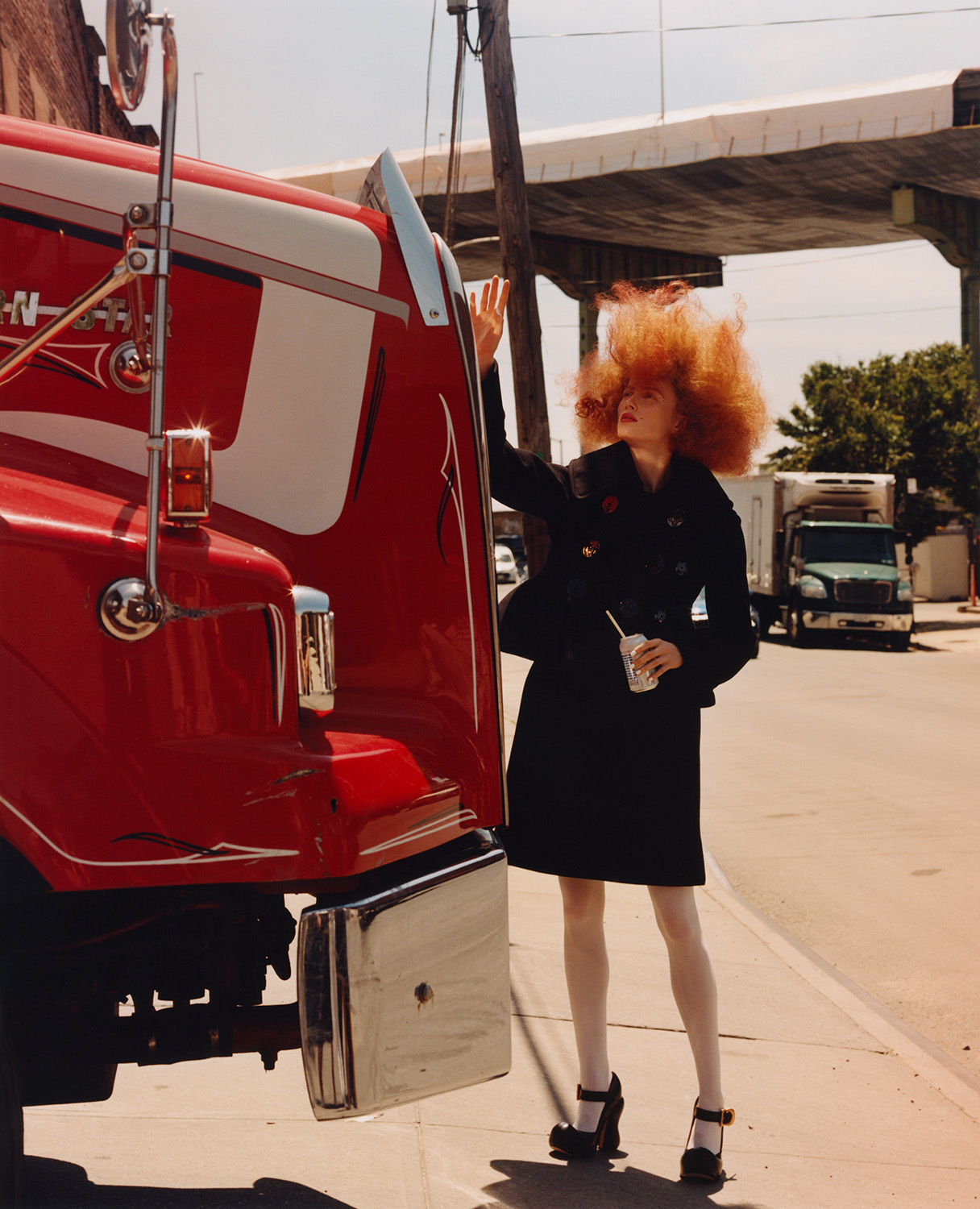 A woman with vibrant red hair poses next to a large red semi-truck on a city street. The image features bright daylight and urban setting.