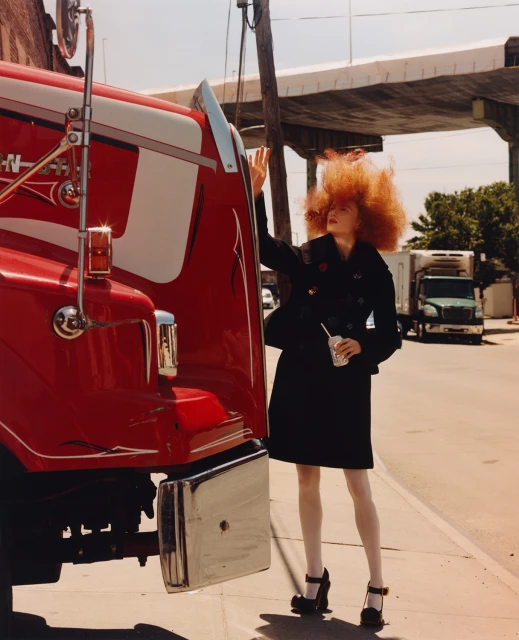 A woman with vibrant red hair poses next to a large red semi-truck on a city street. The image features bright daylight and urban setting.