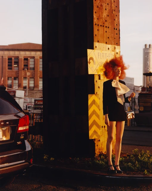 Fashion photography featuring a stylish woman with striking red hair, wearing a black suit with a large bow, standing outdoors near a support beam.