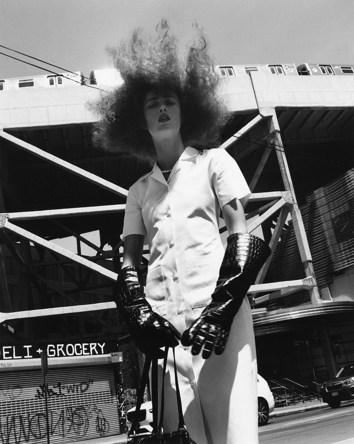 Adult female fashion model wearing oversized black gloves, white shirt, and white pants on an urban street with a raised train.