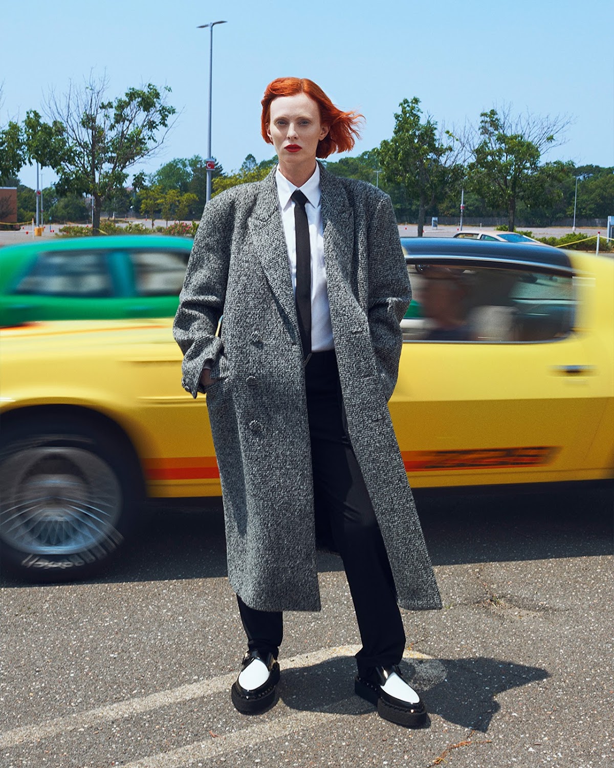 Red-haired woman in a gray overcoat, white button-down shirt, and black tie stands in a parking lot with a yellow car in motion.