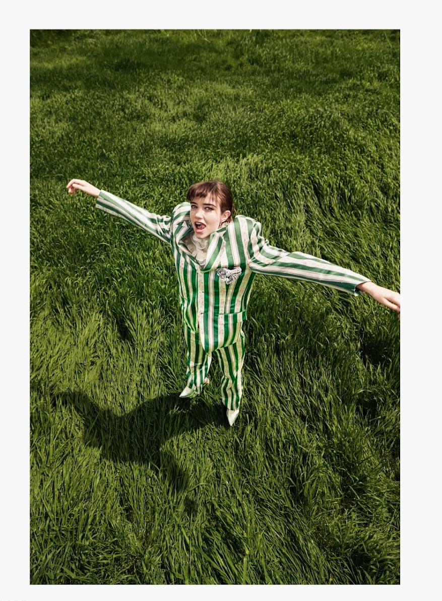High-angle shot of a young woman in a striped green and white suit with arms outstretched in a grassy field, fashion portrait.