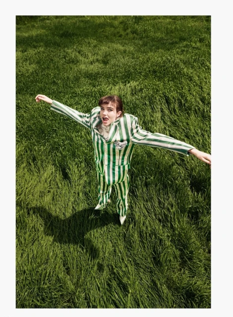 High-angle shot of a young woman in a striped green and white suit with arms outstretched in a grassy field, fashion portrait.
