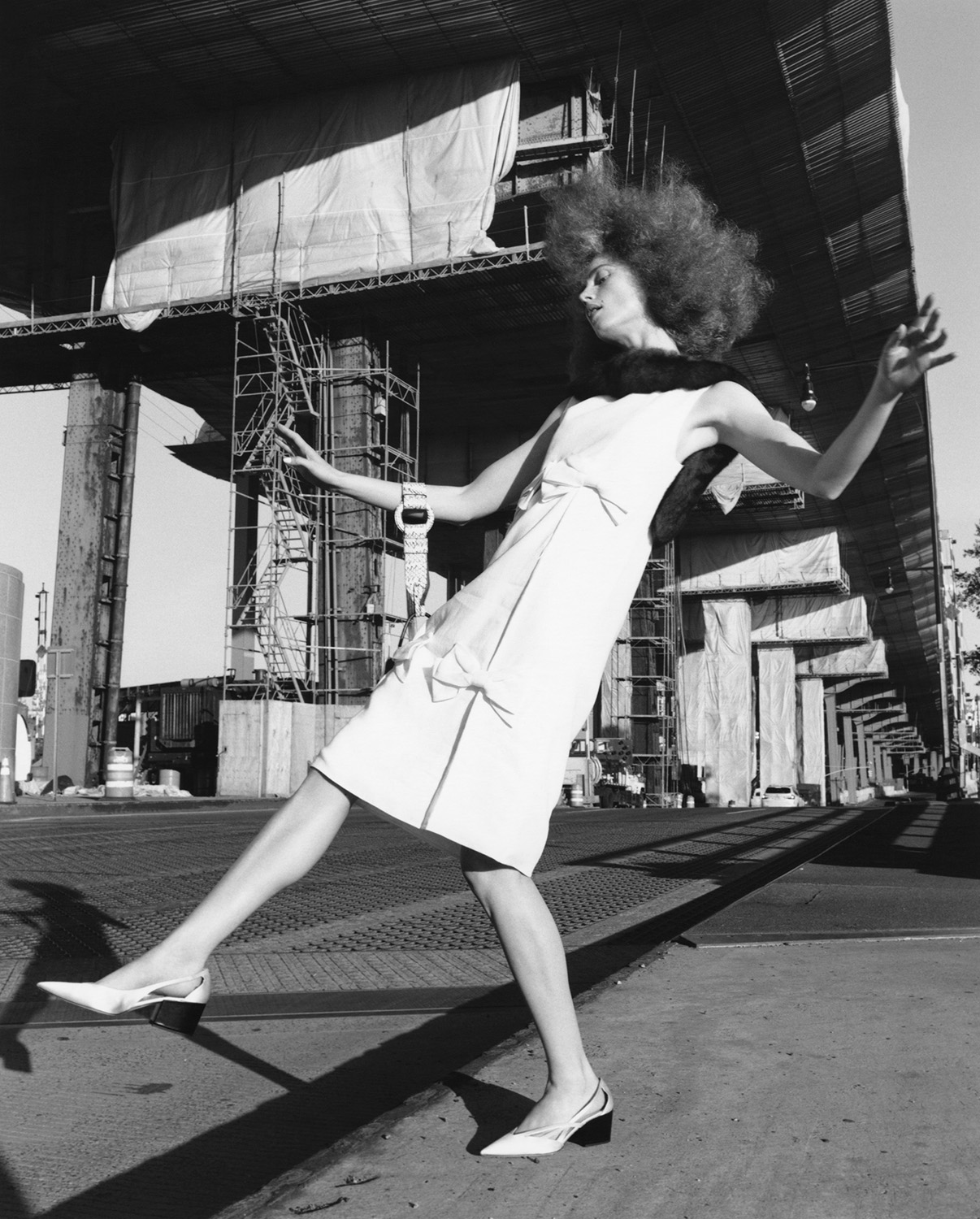 A fashion-forward model with a voluminous afro strikes a pose outdoors in a stylish white dress and slingback heels against an industrial background.