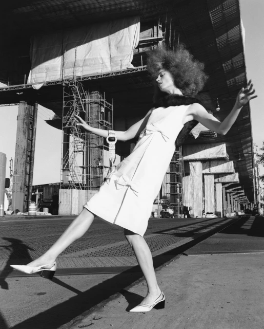 A fashion-forward model with a voluminous afro strikes a pose outdoors in a stylish white dress and slingback heels against an industrial background.